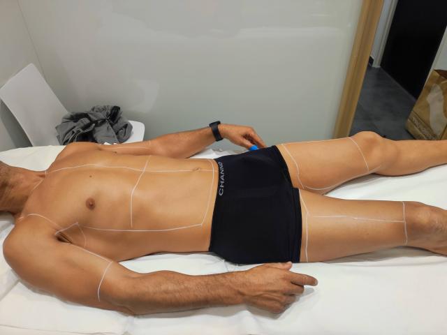 NousCoquins lying on a white table, wearing black underwear, with white lines on their tan, muscular body, pointing with one finger, wearing a black wristband.