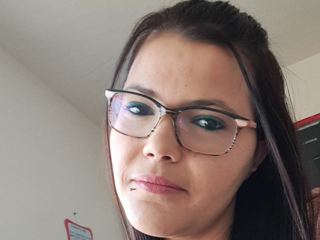 NousCoquins has dark brown hair, wearing glasses with tortoiseshell frames, blue eyeshadow, and a slight pout with a small lip piercing, looking serious.