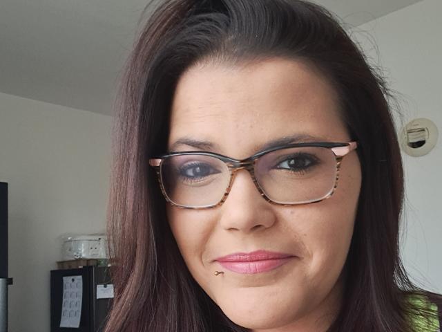 NousCoquins has dark brown hair, glasses with a tortoiseshell pattern, and a small dermal piercing below her pink-lipped right upper lip. Her skin is light with a slight tan.