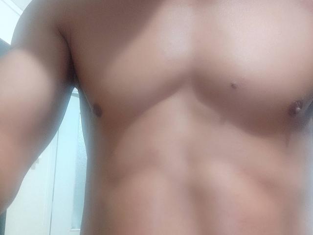 NousCoquins' chest with a smooth, tan, muscular upper body, defined abs, small nipples, and a slight shadow on the left side of the image.