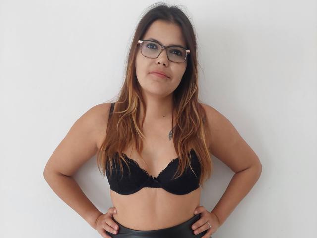 NousCoquins with glasses, brown hair, black bra, hands on hips, looking at camera, slight nose piercing, white wall background, standing, showing small tits.