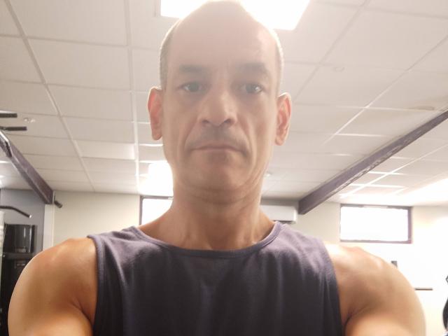 NousCoquins has a bald head, muscular arms, and is wearing a sleeveless dark blue shirt, looking down at the camera with a neutral expression in a brightly lit room.