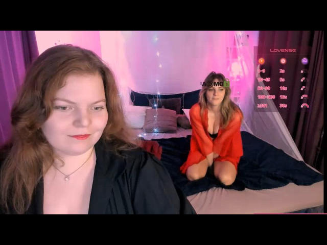 Two JoseReneSmee figures in a bedroom. Foreground JoseReneSmee has wavy brown hair, pale skin, red lipstick, and a black robe. Background JoseReneSmee, with similar hair and makeup, kneels on a bed wearing an open red robe, revealing a black bra and cleavage. The bed has dark blue and beige bedding, with pink and purple lighting. A digital overlay with red text and numbers is on the right. The overall composition has a soft, intimate feel.