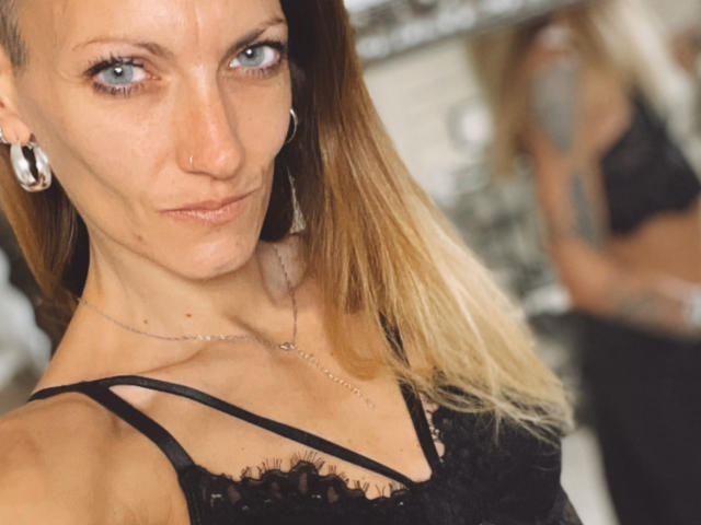 DaneeSoHot hat blondes Haar, blaue Augen und eine Nasenring. Sie trägt eine schwarze Spitzenbluse, die ihre kleinen Brüste zeigt, und im Hintergrund ist eine unscharfe Reflexion ihres Gesäßes zu sehen..