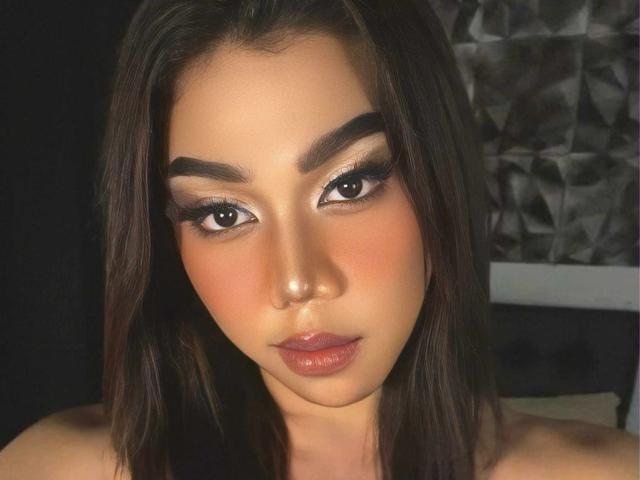 FelizOganda tiene el cabello castaño oscuro, cejas pobladas, delineador de ojos negro brillante y lápiz labial rosado-rojo. Está mirando directamente a la cámara con una expresión neutra. Sus senos no son visibles..