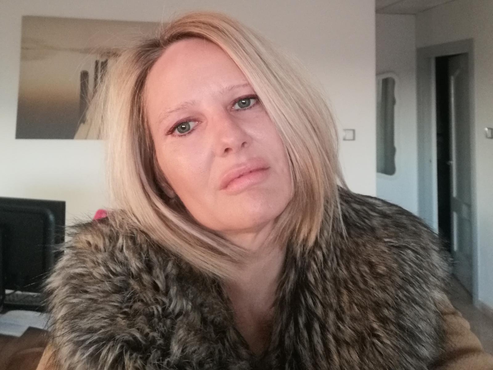 NatjeStout with blonde hair, wearing a fur-lined coat, has a neutral expression, light skin, and blue eyes. Her ass is not visible, just her face and coat.