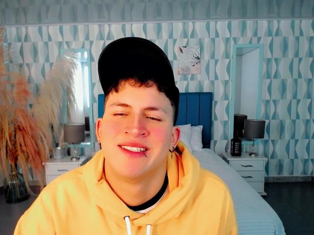 AxelSavage, un homme corpulent avec des cheveux noirs bouclés, portant une casquette noire et un sweat à capuche jaune, souriant avec un large sourire édenté, les yeux fermés, dans une chambre très lumineuse..