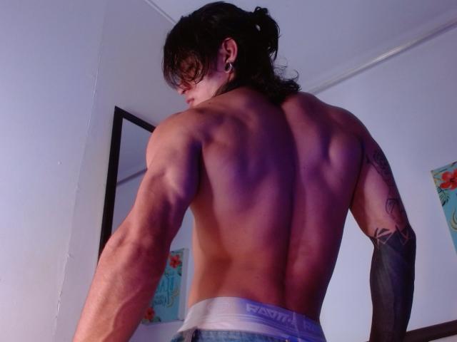 AlexBernal, ohne Hemd, muskulöser Rücken, definierte Muskeln, Tattoos am Arm, dunkles Haar im Pferdeschwanz, Ohrringe, weiße Unterwäsche, stehend, Zimmer mit Postern, niedriger Kamerawinkel, gedämpfte Beleuchtung..