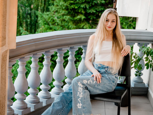 MiaMeowa with long blonde hair, white tube top, and blue jeans with white graffiti, sitting on black chair, greenery outside, smiling, hand on chair.