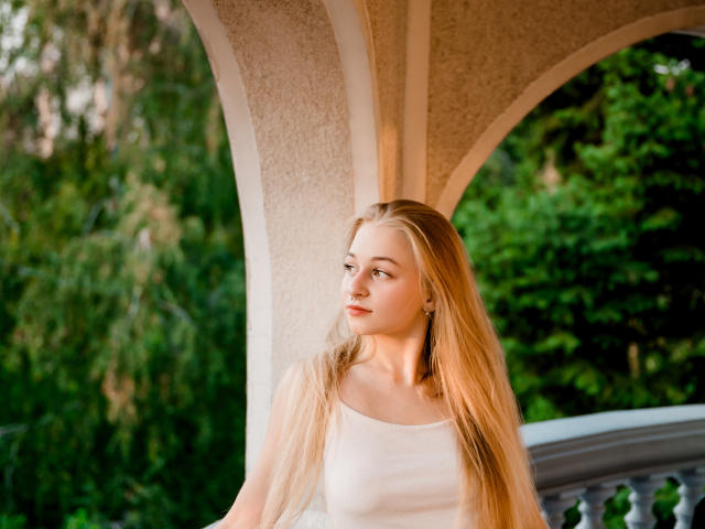 MiaMeowa with long blonde hair, small tits, and a cute face, wearing a white tank top, standing under an arch, looking to the side with a slight smile.