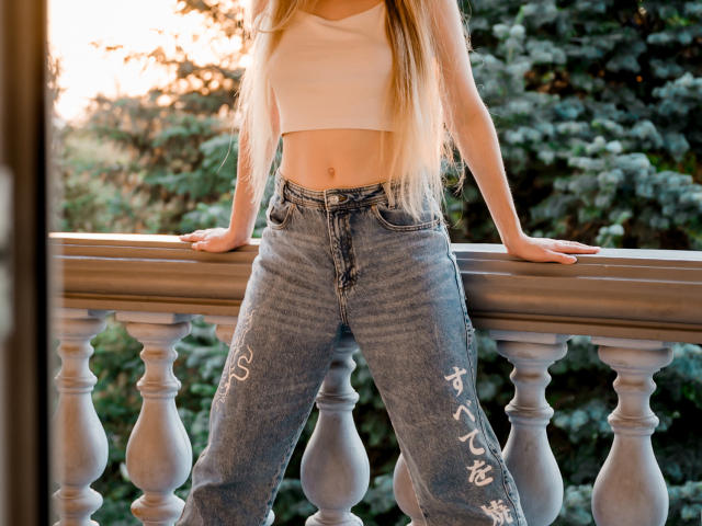 MiaMeowa, avec de longs cheveux blonds, un débardeur blanc et un jean bleu avec des caractères japonais blancs sur les jambes, appuyée sur la rambarde d'un balcon, les mains sur la rambarde..
