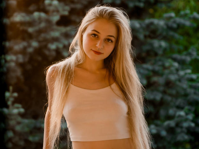 MiaMeowa with long blonde hair, small boobs, wearing a white crop top, smiling gently, standing in a forest, sunlight filtering through trees.