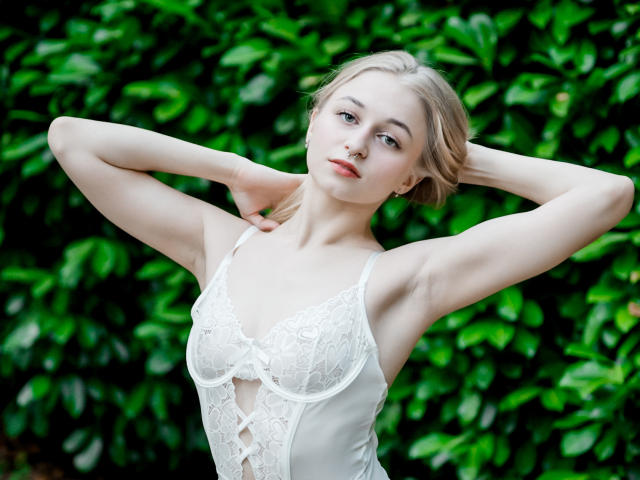 MiaMeowa, les bras levés, portant un haut en dentelle blanche, montrant de petites seins, une peau pâle, des cheveux blonds et un visage sérieux, sur un fond de feuilles vertes..