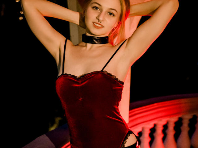 MiaMeowa with arms raised, wearing a red velvet bodysuit with black lace, black choker, and black straps, smiling, showing her tits, in dim, red-lit setting.