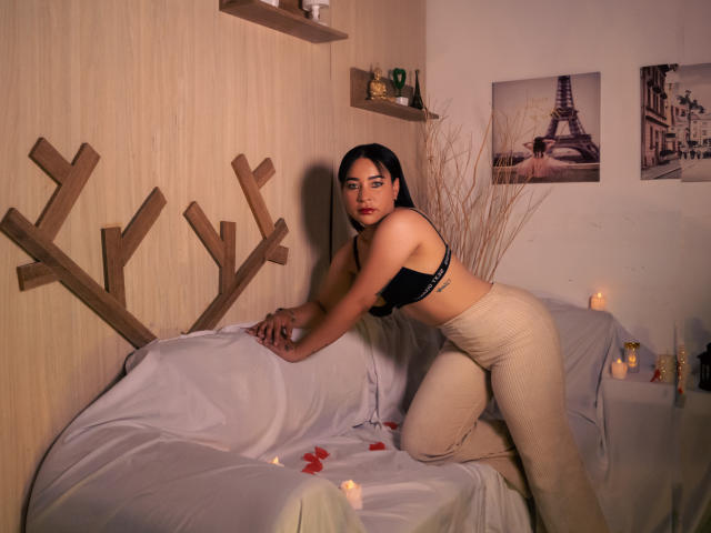 SaharaArisa with dark hair, wearing a black bra and beige pants, on a white-covered couch, leaning forward, candles, rose petals, wooden wall art, and Paris photos in the background.