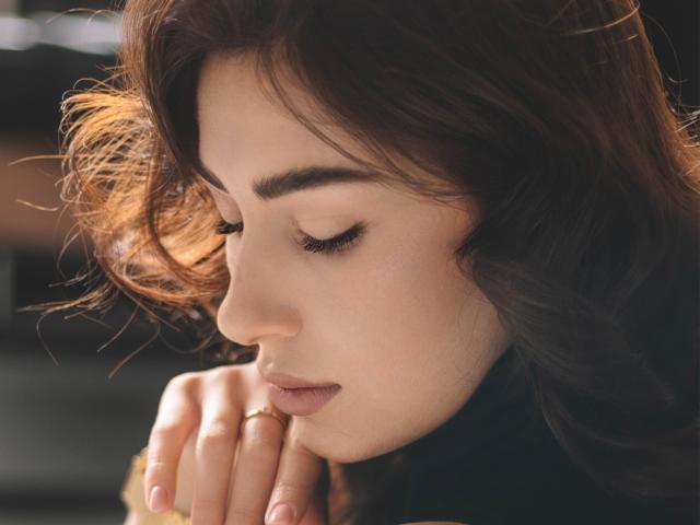 OliviaCluff's face with wavy brown hair, closed eyes, and soft pink lips, gently resting her hand near her mouth, showing a calm and peaceful expression.