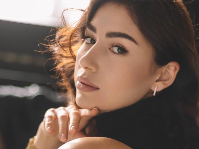 OliviaCluff with wavy brown hair, subtle makeup, and pink lipstick, looking over her shoulder with a gentle smile, holding her chin, wearing small white earrings.