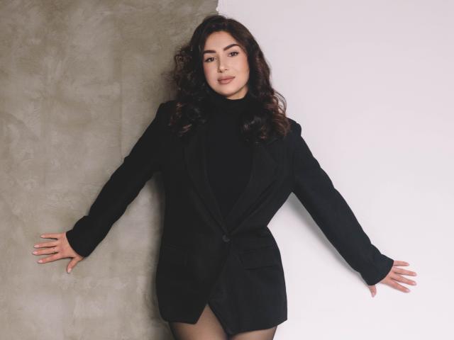 OliviaCluff with curly brown hair, black turtleneck, and black blazer, arms spread, standing against a gray and white wall, looking confident, smiling slightly, showing a cute ass.