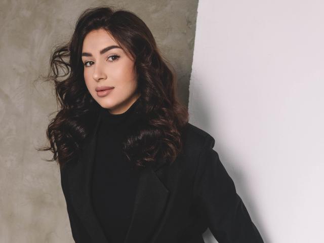 OliviaCluff with wavy dark brown hair, wearing a black turtleneck and black blazer, looking slightly to the side with a subtle smile, standing against a white wall.