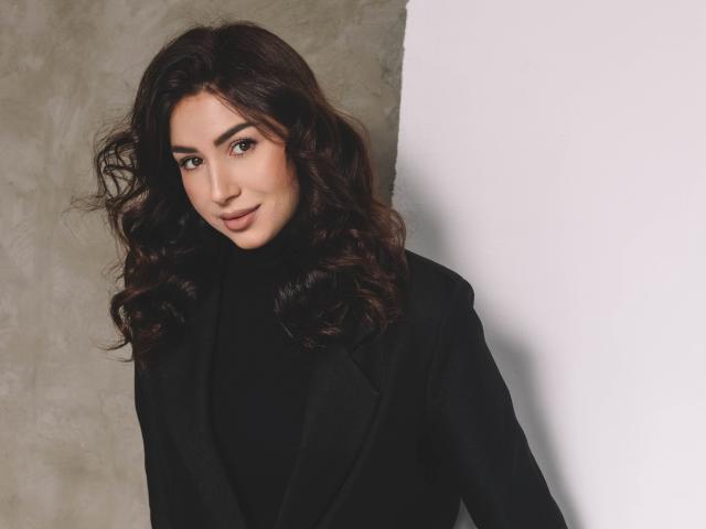 OliviaCluff with wavy dark brown hair, wearing a black turtleneck and black blazer, smiling subtly. Her brown eyes are softly looking at the camera.