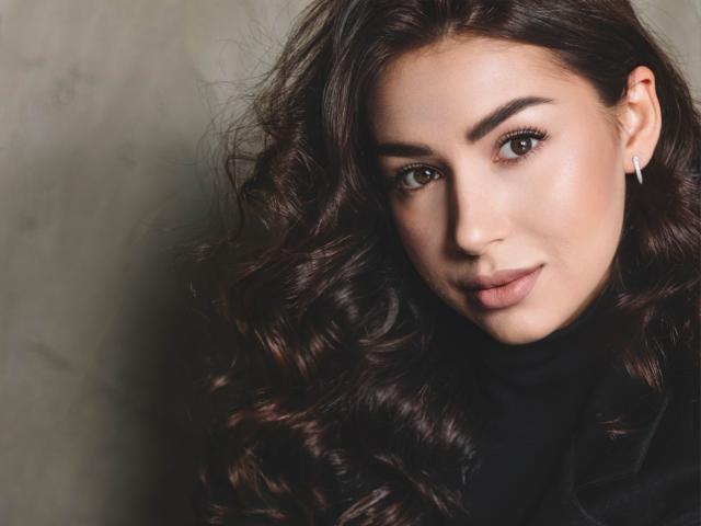 Olivia Cluff heeft lang, golvend bruin haar, een lichte huid en een schattige glimlach met subtiele make-up. Ze draagt een zwarte coltrui en kleine witte oorbellen en kijkt recht in de camera..