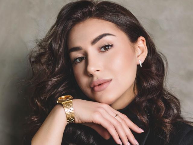 OliviaCluff has wavy brown hair, green eyes, and a subtle smile. She's wearing a gold wristwatch and diamond earrings, with her hand resting on her chin.
