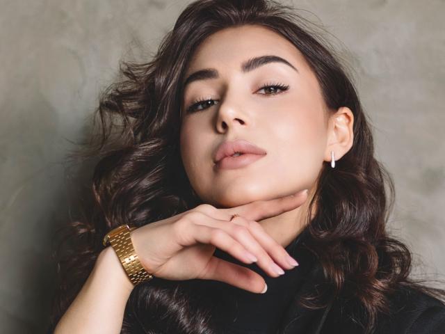 OliviaCluff has wavy brown hair, brown eyes, and is looking up with a slight smile. She's wearing a gold watch and a pearl earring, with her hand near her face.