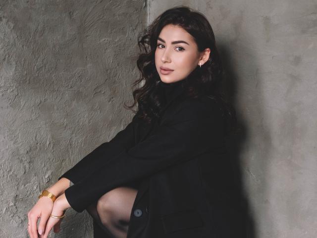 OliviaCluff with curly dark hair, wearing a black coat, squatting against a textured wall, gold wristwatch, subtle makeup, and a small earring, looking slightly to the side.