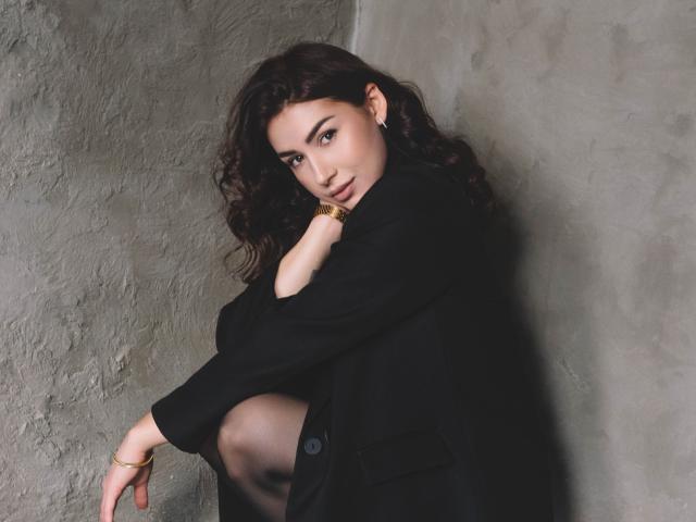 OliviaCluff with curly brown hair, black coat, and gold bracelet, crouching against a rough gray wall, looking over her shoulder with a subtle smile, showing her taut ass.
