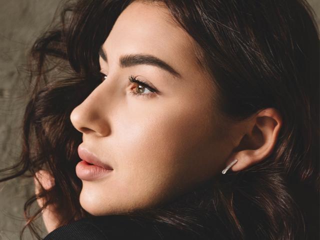 OliviaCluff's face with wavy brown hair, brown eyes, and full lips, showing a slight smile, wearing a single stud earring in her right ear. Smooth, sexy, and attractive.