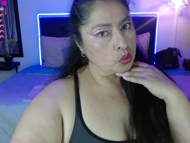 Keyla69Hot has dark hair, pink eyeliner, red lipstick, and gold earrings. She's pouting with her hand near her mouth, wearing a black tank top, showing her tits.