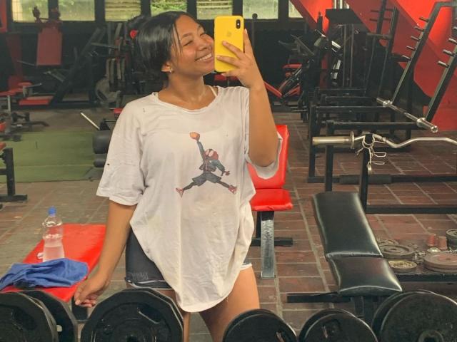 DreamingOfU with dark hair, smiling, wearing a white T-shirt with a cartoon, holding a yellow phone, in a gym, standing near weights and a red bench.