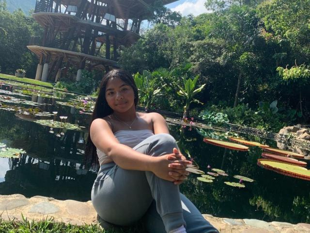DreamingOfU with long black hair, small tits, and a cute smile, sitting by a lily pad-filled pond, wearing a white strapless top and gray pants, hands on knees.