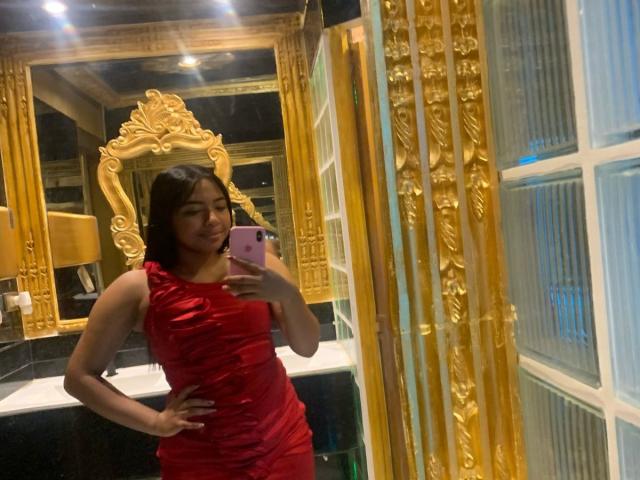 DreamingOfU in a red dress, holding a pink phone, posing in front of a gold-framed mirror, with a hand on her hip, smiling slightly.