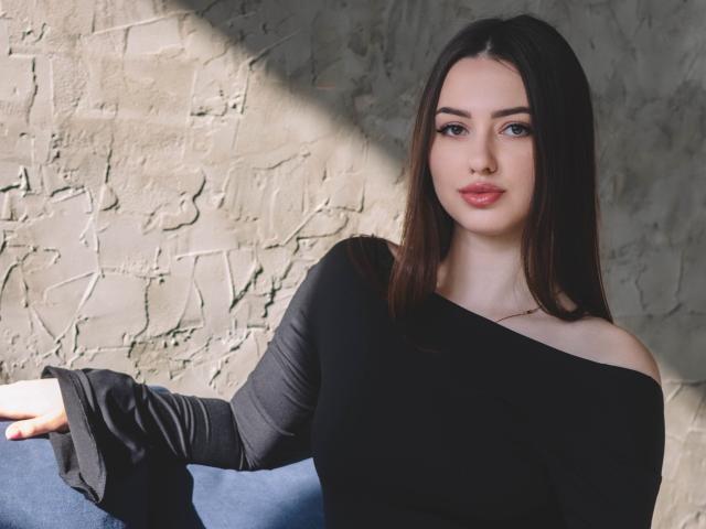 ChristinaBliss with long brown hair, light skin, and pink lips, wearing a black off-shoulder top, sitting on a blue surface, her right arm resting on it.