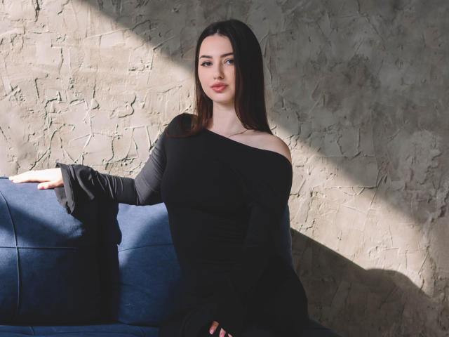 ChristinaBliss, con cabello largo y oscuro, vestida con un vestido negro de corte asimétrico, sentada en un sofá azul, con una sombra suave, pared con textura, mano derecha sobre el sofá, brazo izquierdo relajado, sonrisa sutil, piel clara..