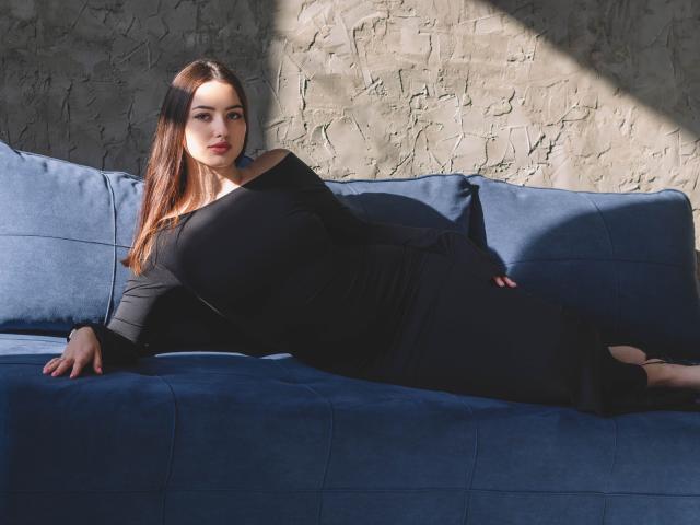 ChristinaBliss, con cabello castaño largo, acostada en un sofá azul, vestida con un vestido negro de corte asimétrico que deja un hombro al descubierto, mostrando su gran trasero y sus pequeños senos, a la luz del sol, mirando a la cámara..
