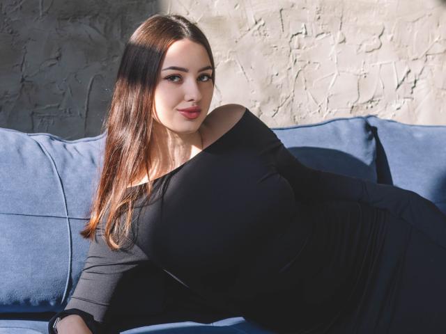 ChristinaBliss with big tits, lying on a blue couch, wearing a black off-shoulder dress, looking at the camera with a slight smile, sunlight on her face.