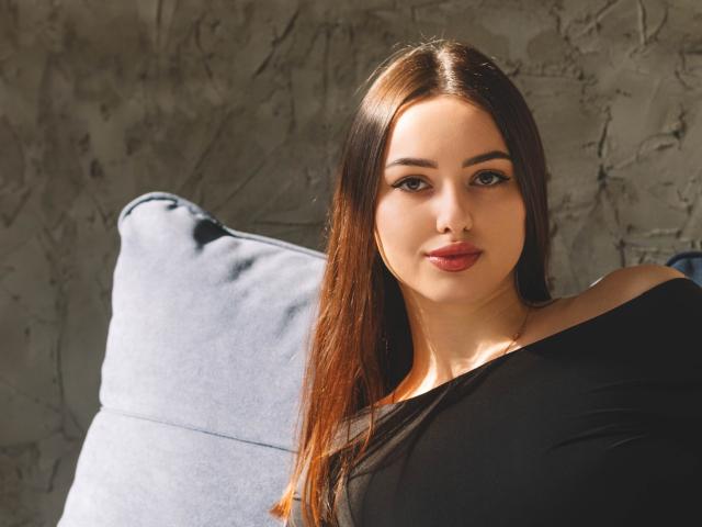 ChristinaBliss has long, straight brown hair with subtle highlights, parted in the middle. She's wearing a black off-shoulder top that reveals a bit of her collarbone. Her makeup is subtle with dark eyeliner and pink lipstick. She's sitting on a blue, textured couch with a gray, rough-textured wall in the background. The light is coming from the left, creating a natural shadow on the right side of her face. Her expression is calm and slightly smiling. The image is well-lit and sharp.