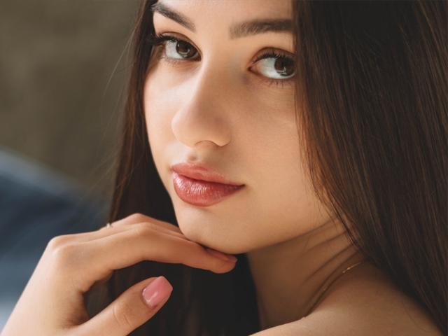 Close-up of ChristinaBliss's face. She has long, straight brown hair, slightly parted lips with glossy pink lipstick, and brown eyes looking to the side. Her right hand is near her chin, with pink-painted nails. Her skin is smooth and light, with subtle makeup highlighting her natural features. The image is sharply focused on her face, with a soft, warm light enhancing her skin's texture. The background is blurred, keeping the focus on ChristinaBliss. The composition is centered, emphasizing her facial features and expression.