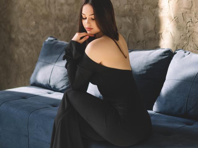 ChristinaBliss, com cabelos longos e escuros, vestindo um vestido preto com um ombro descoberto, sentada em um sofá azul, com os seios levemente visíveis, a mão perto do rosto, luz solar suave, parede cinza texturizada ao fundo..