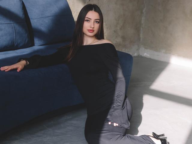 ChristinaBliss, con cabello castaño largo, lápiz labial rojo, una blusa negra con un hombro descubierto, pantalones grises y tacones negros, apoyada en un sofá azul, sentada en el suelo de concreto, con la luz del sol proyectando sombras..