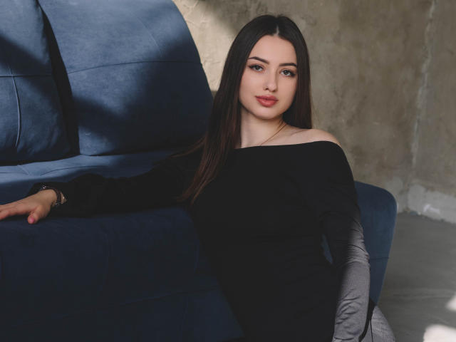 ChristinaBliss with long black hair, off-shoulder black top, sitting on blue couch, right arm on cushion, gray sleeve, red lips, subtle makeup, relaxed, looking at camera.