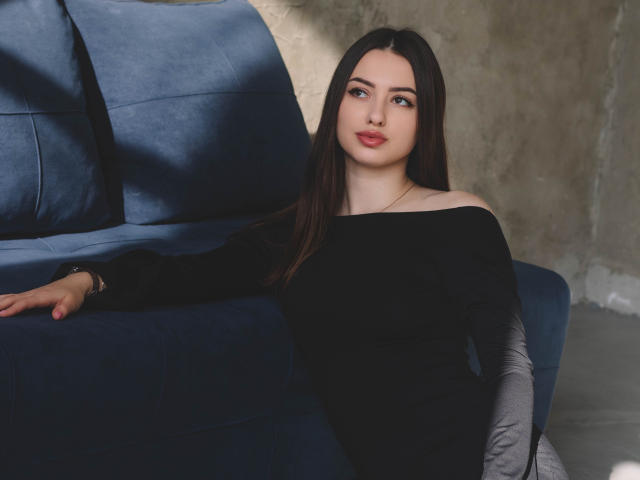 ChristinaBliss is a young woman with fair skin and long, straight brown hair. She wears a black off-shoulder long-sleeve top. Her blue eyes and red lipstick stand out. She leans against a blue velvet sofa with her right arm resting on the back. The textured beige wall is in the background. She has a subtle, confident expression. The image uses soft, natural lighting. She wears a thin gold necklace. The composition is centered and balanced.