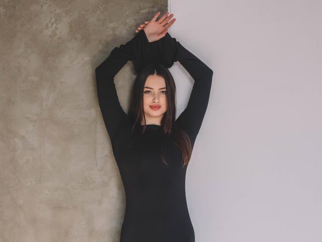 ChristinaBliss heeft donkerbruin haar, draagt een zwarte top met lange mouwen, haar armen zijn omhoog, haar handen raken de muur, ze glimlacht, heeft bleke huid, kleine borsten en staat tegen een muur die grijs en wit is..
