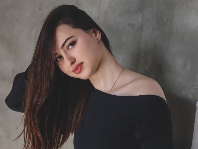 ChristinaBliss with long brown hair, off-shoulder black top, pale skin, subtle makeup, red lips, slight smile, hand in hair, delicate gold necklace, soft light, confident look.