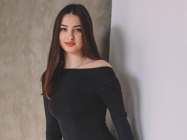 ChristinaBliss with long brown hair, wearing a black off-shoulder top, showing her tits, standing against a white wall, smiling with red lipstick.