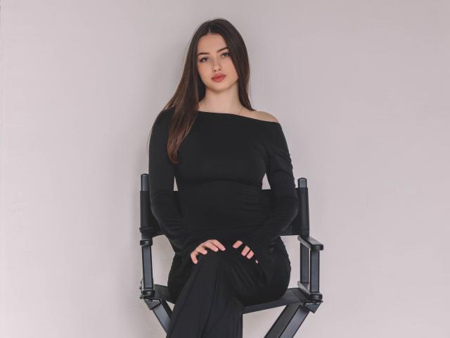 ChristinaBliss with long brown hair, off-shoulder black dress, sitting on a director's chair, hands on knees, looking at camera, pale skin, red lipstick, neutral expression.