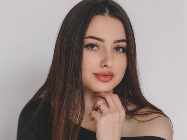 ChristinaBliss has long brown hair, green eyes, and glossy pink lips. She's looking at the camera with a slight smile, her hand touching her chin. She's wearing a black top.