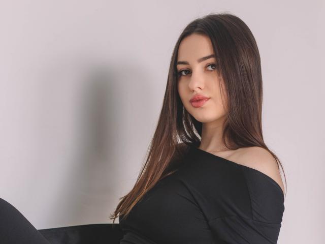 ChristinaBliss with long brown hair, black off-shoulder top, and pouty lips, looking directly at the camera, showing her tits slightly, against a plain white background.